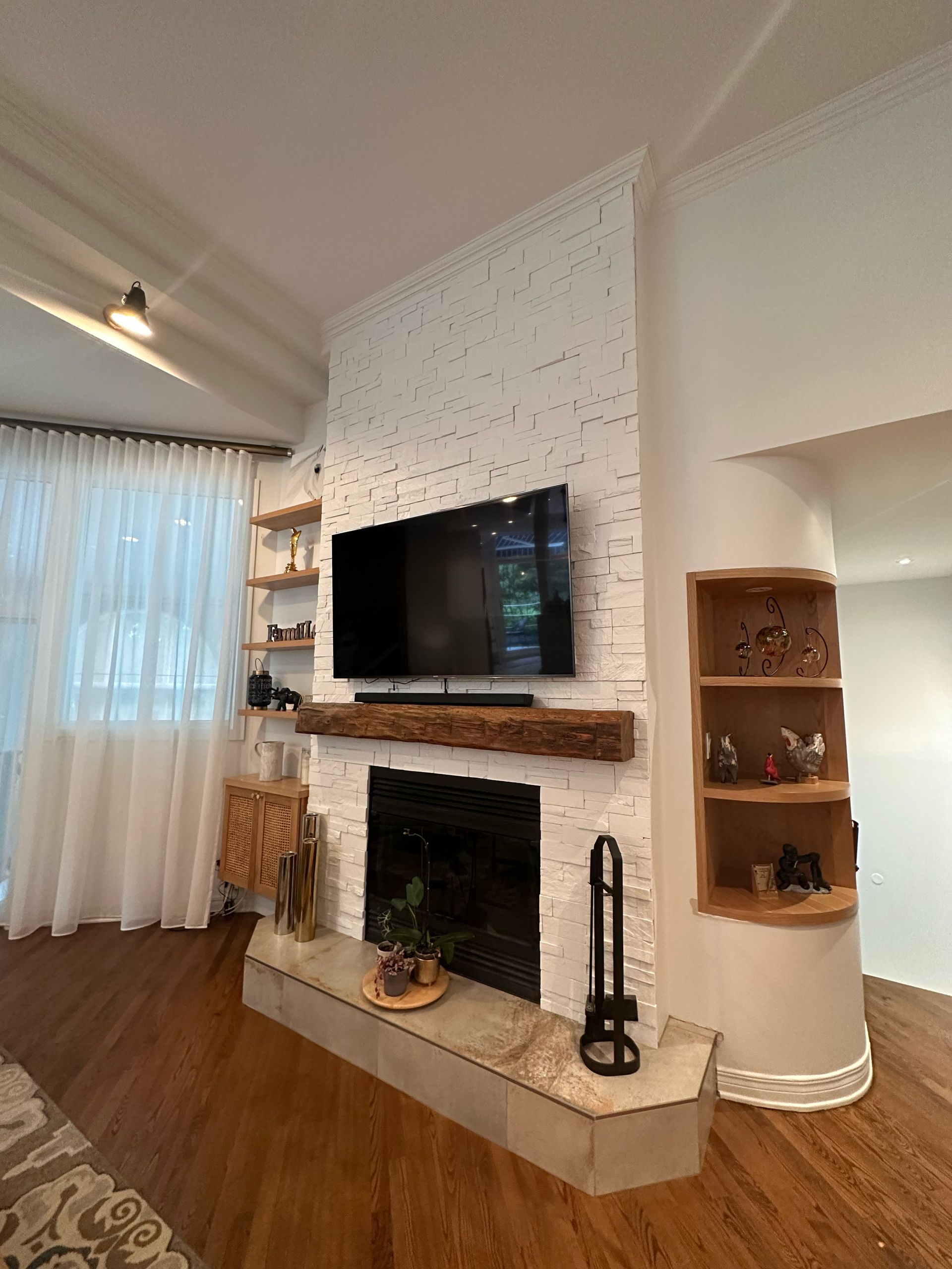 Salon avec cheminée en briques blanches, télévision, étagères en bois et une fenêtre avec des rideaux transparents.