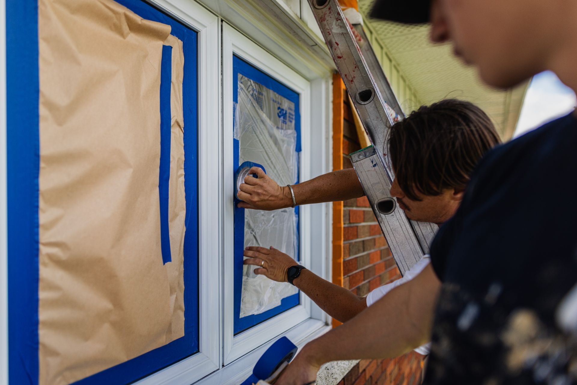 Deux personnes peignent une fenêtre avec du ruban adhésif bleu.