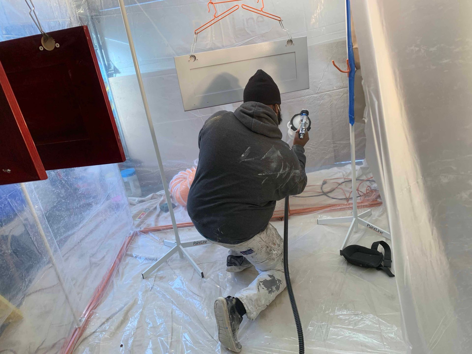 Un homme peint à la bombe dans une cabine de peinture transparente. Il porte un sweat à capuche, est agenouillé et tient un manomètre.