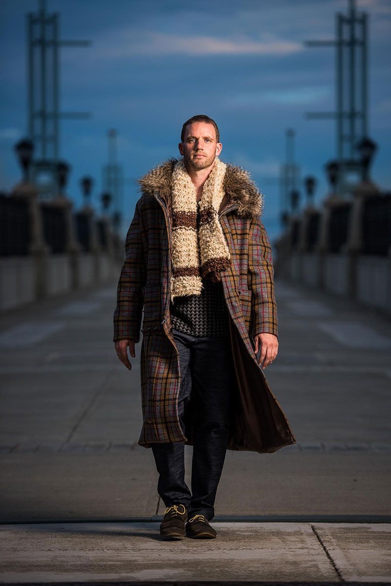 Man in long plaid coat and fur scarf walks on a bridge at dusk.