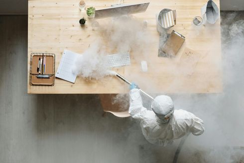 A man in a protective suit is disinfecting a desk with a sprayer.