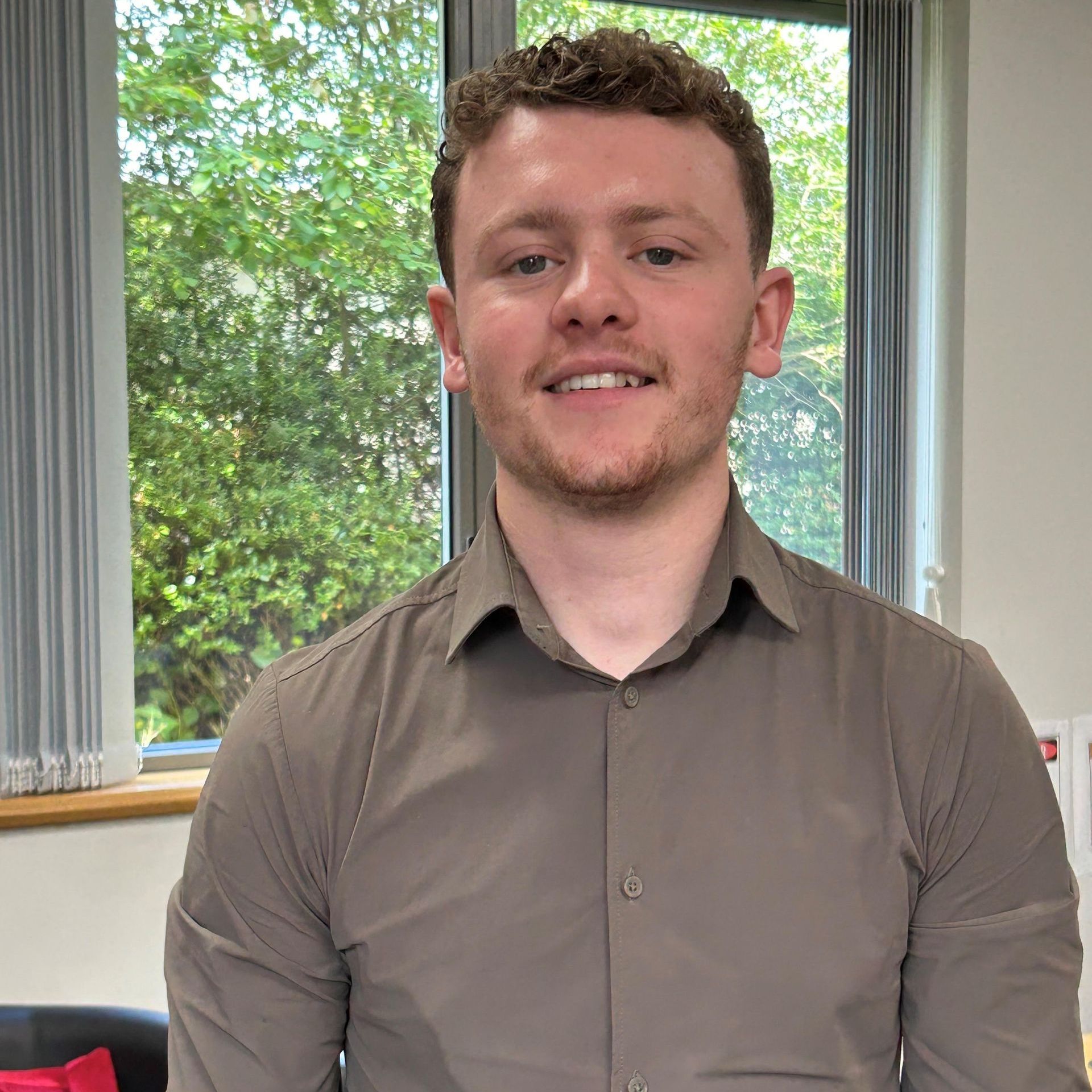 A man in a grey shirt is smiling in front of a window.