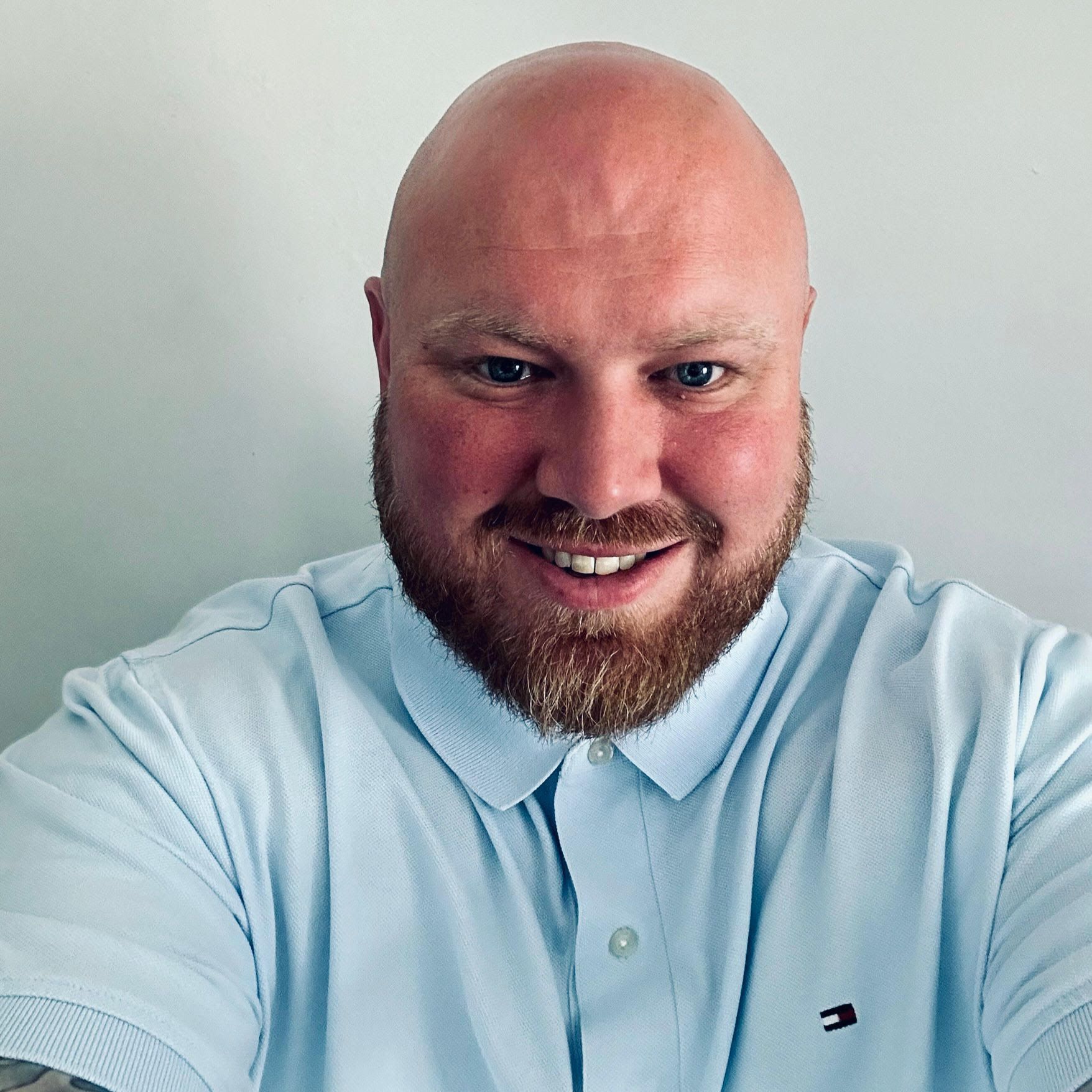 A bald man with a beard is wearing a light blue polo shirt.