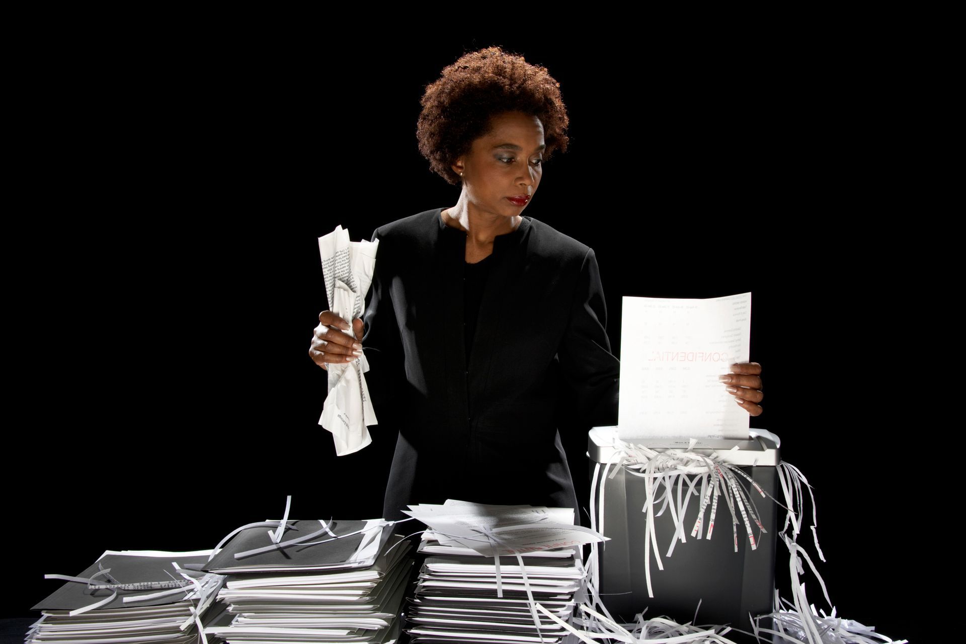 Woman performing paper shredding services with stacks of paper and a shredder in dark office. Woman performing paper shredding services with stacks of paper and a shredder in dark office.
