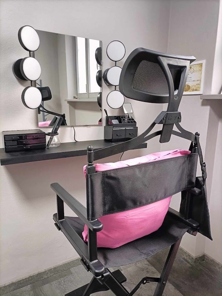 Postazione trucco con sedia da regista nera, specchio e luci. Cuscino rosa visibile.