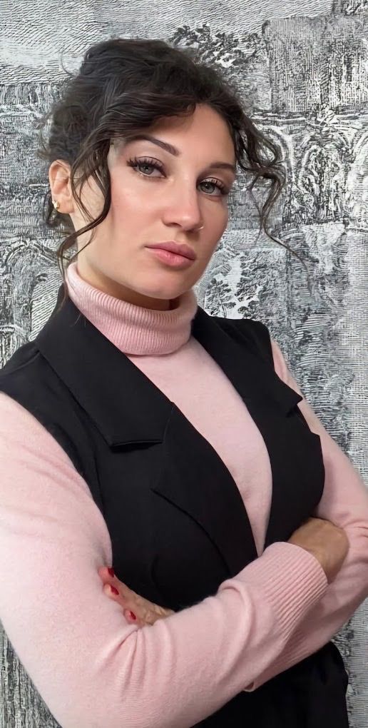 Donna con le braccia incrociate, che indossa un gilet nero e un dolcevita rosa, in posa davanti a una parete decorata.