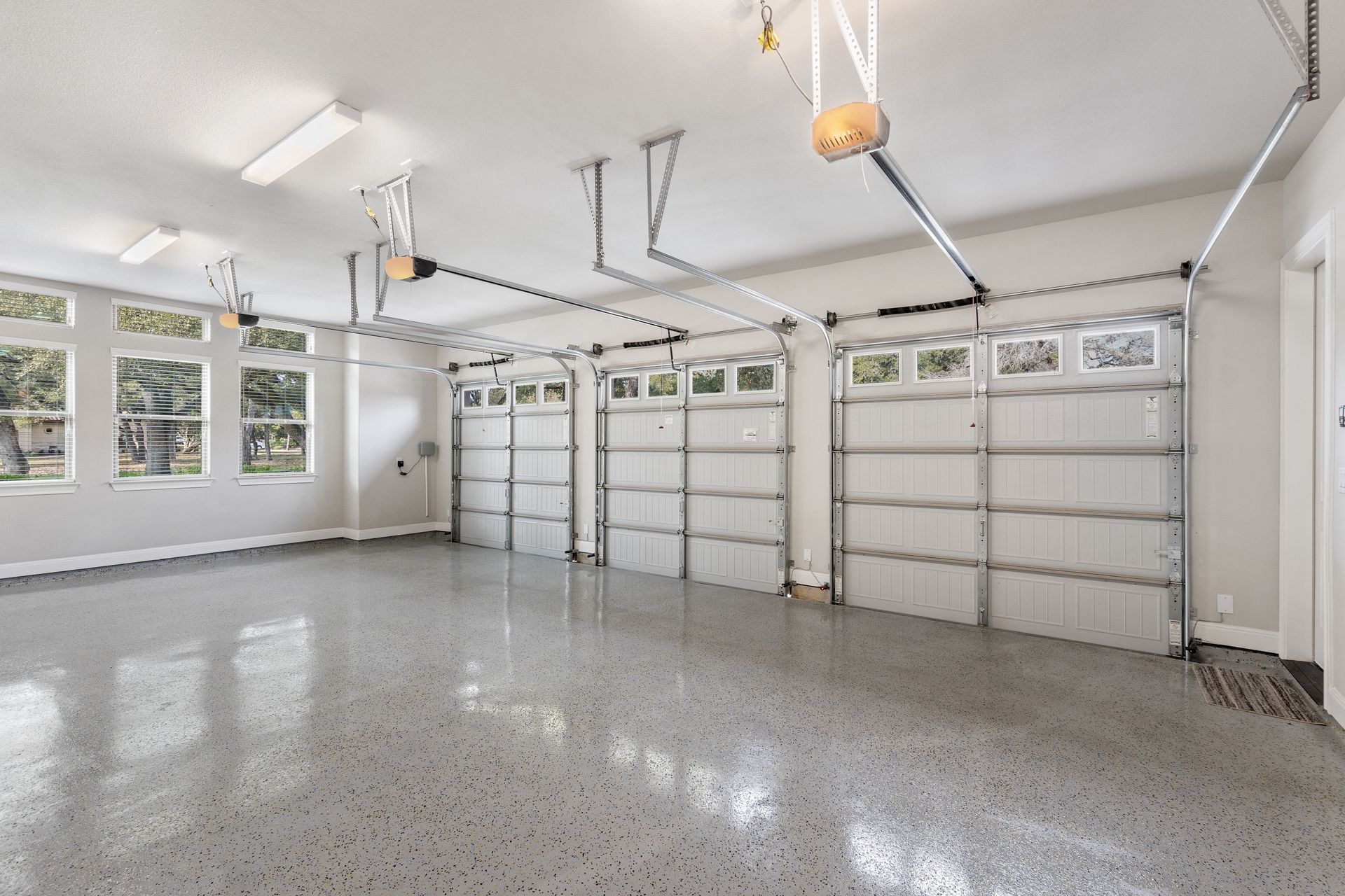 Empty, spacious garage with three closed white doors, speckled floor, and windows.