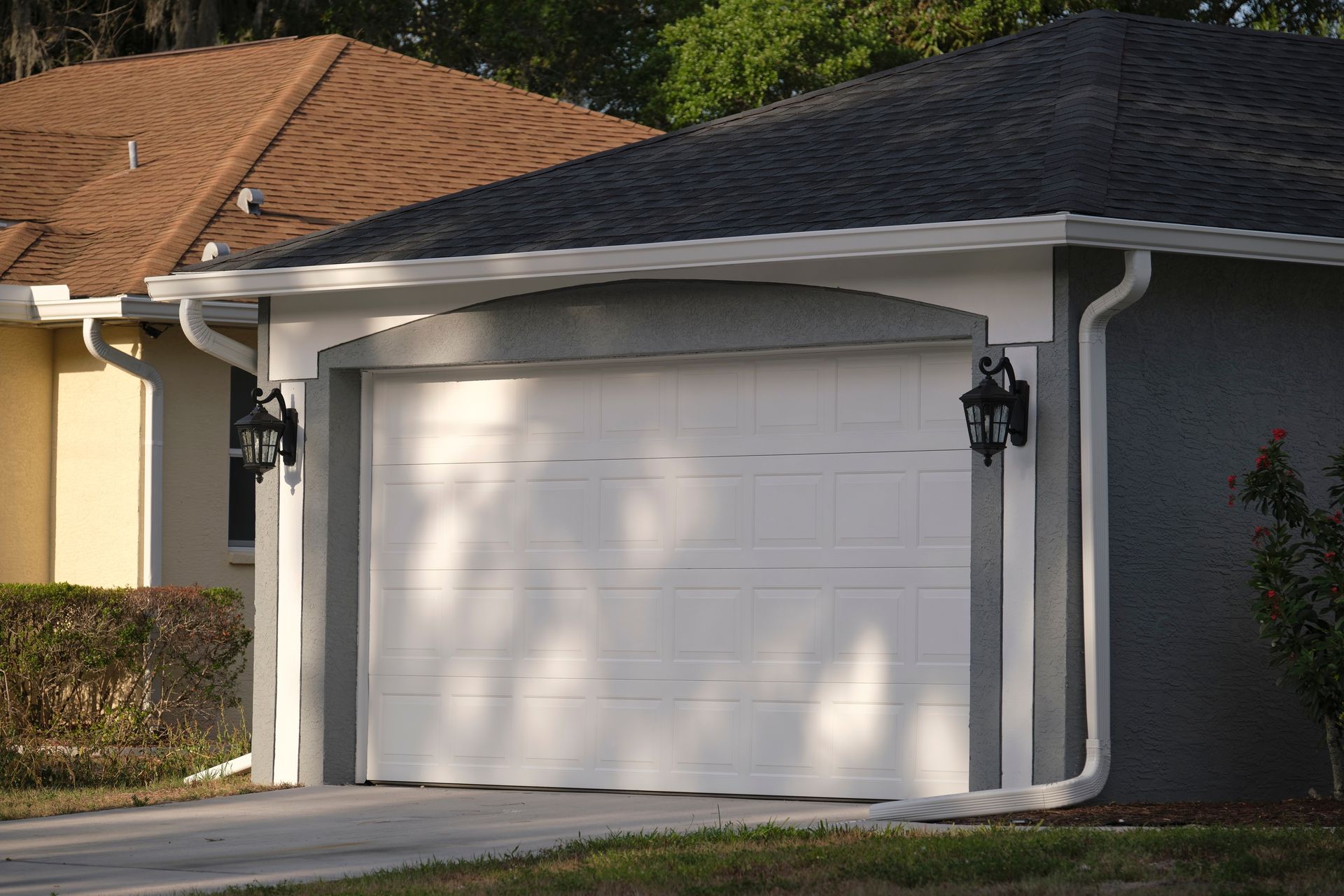 White garage door on a gray house with white trim, black lamps, and brown roof behind.
