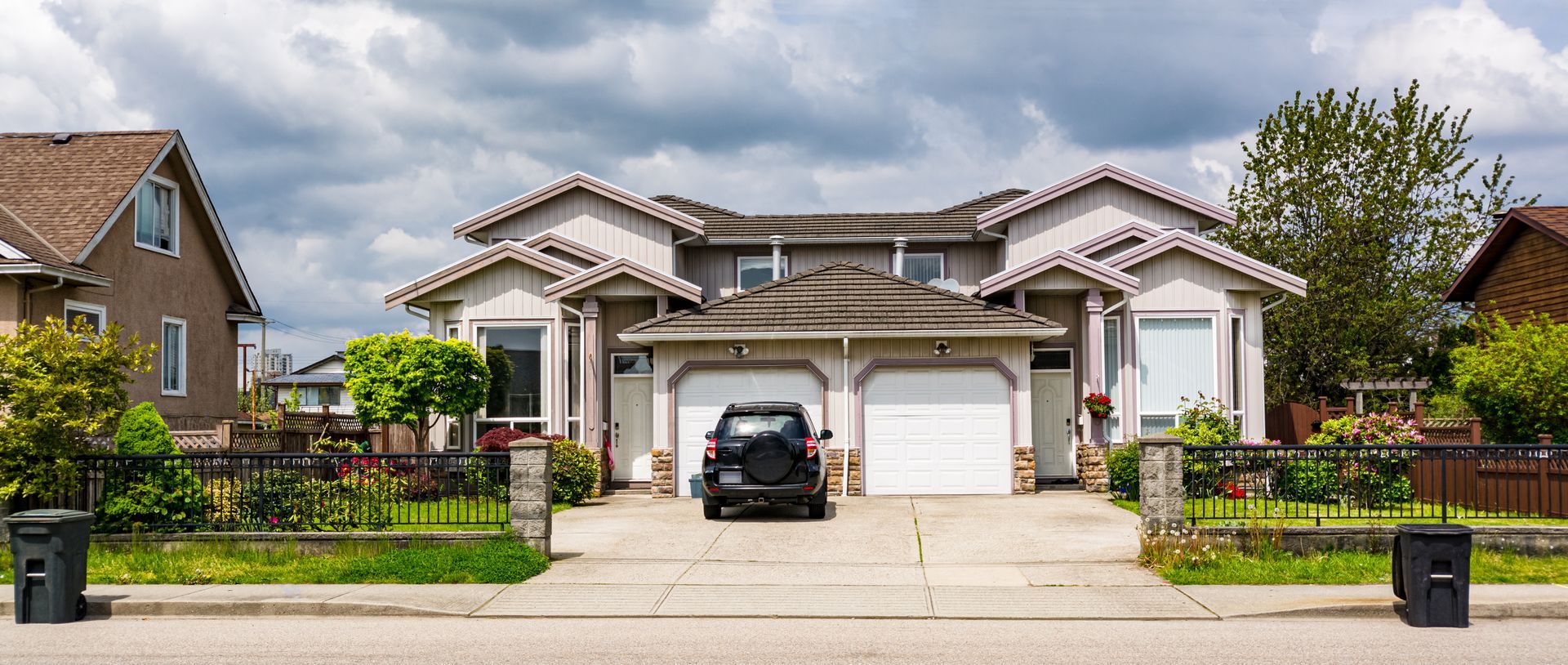 A duplex with two garages and a black SUV parked in the driveway, under a cloudy sky.