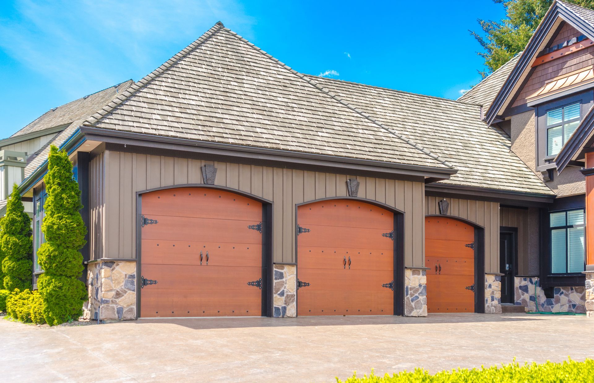 Brown-and-tan garage with three arched doors, stone accents, and shingled roof under a blue sky.