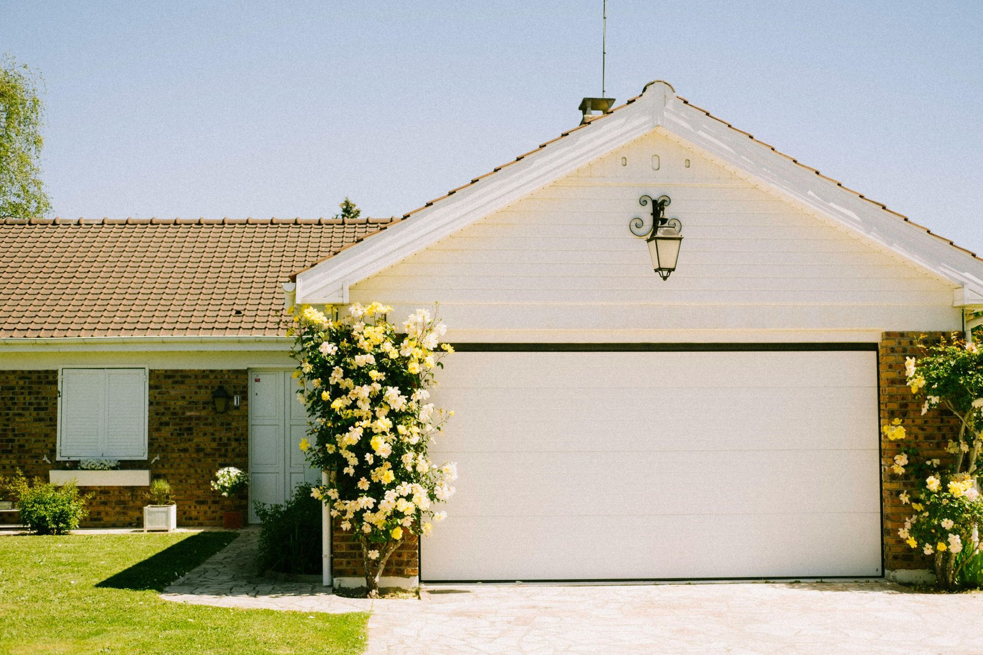 White garage with flowering bush in front of a home with brown roof, blue sky.