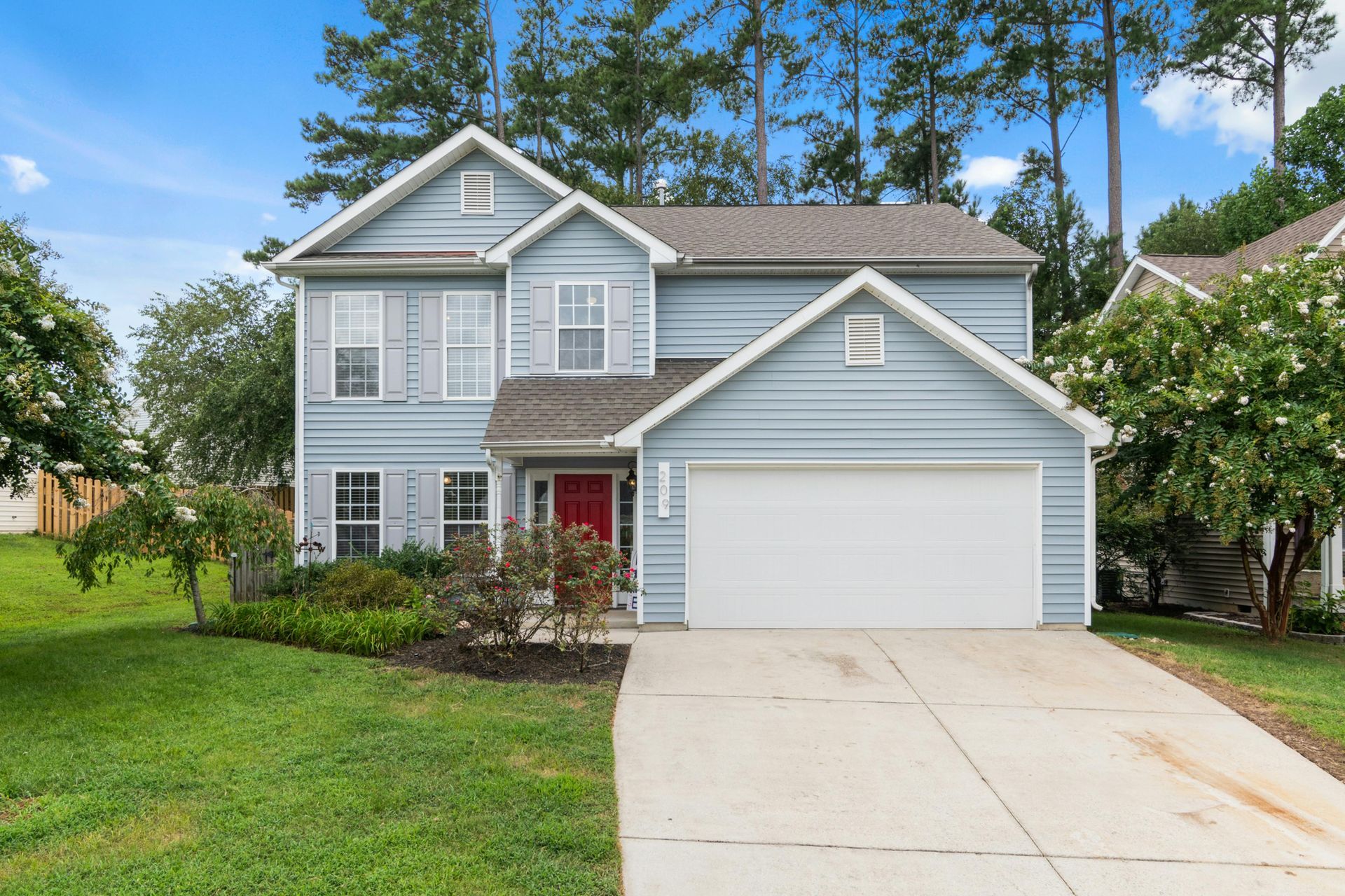 Two-story blue house with white garage door and red front door, set on a green lawn with trees.