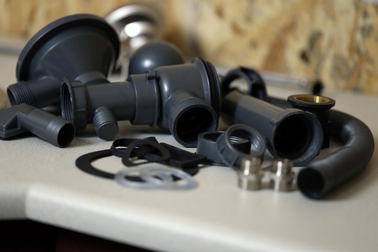 Assorted gray plumbing parts, including pipes, connectors, and washers, sit on a white surface, blurred wood background.