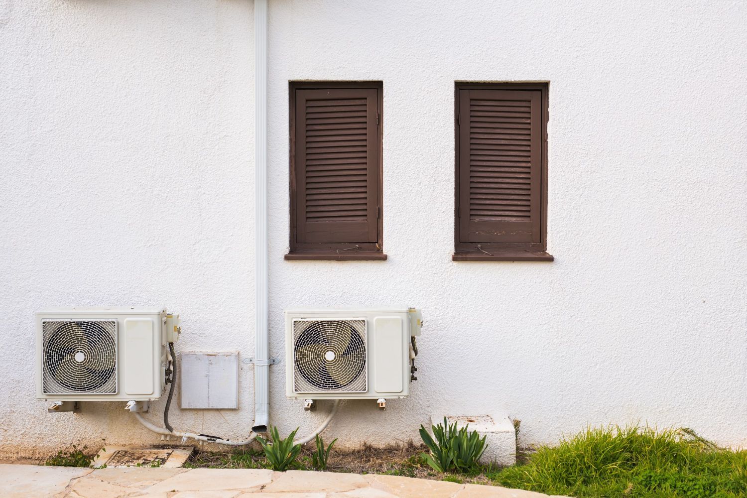 White wall with two brown shuttered windows and two air conditioning units.