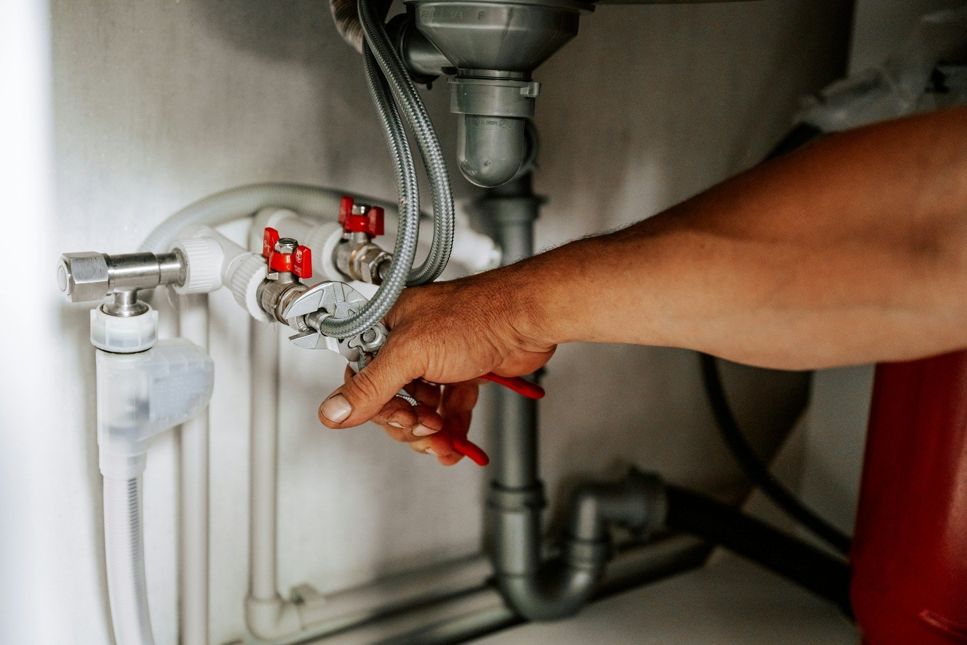 A person's hand turning a red valve on plumbing pipes in a basement.