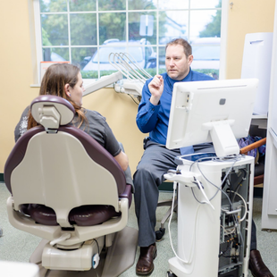 Dentist pointing at a tablet screen while talking with a patient in an office.