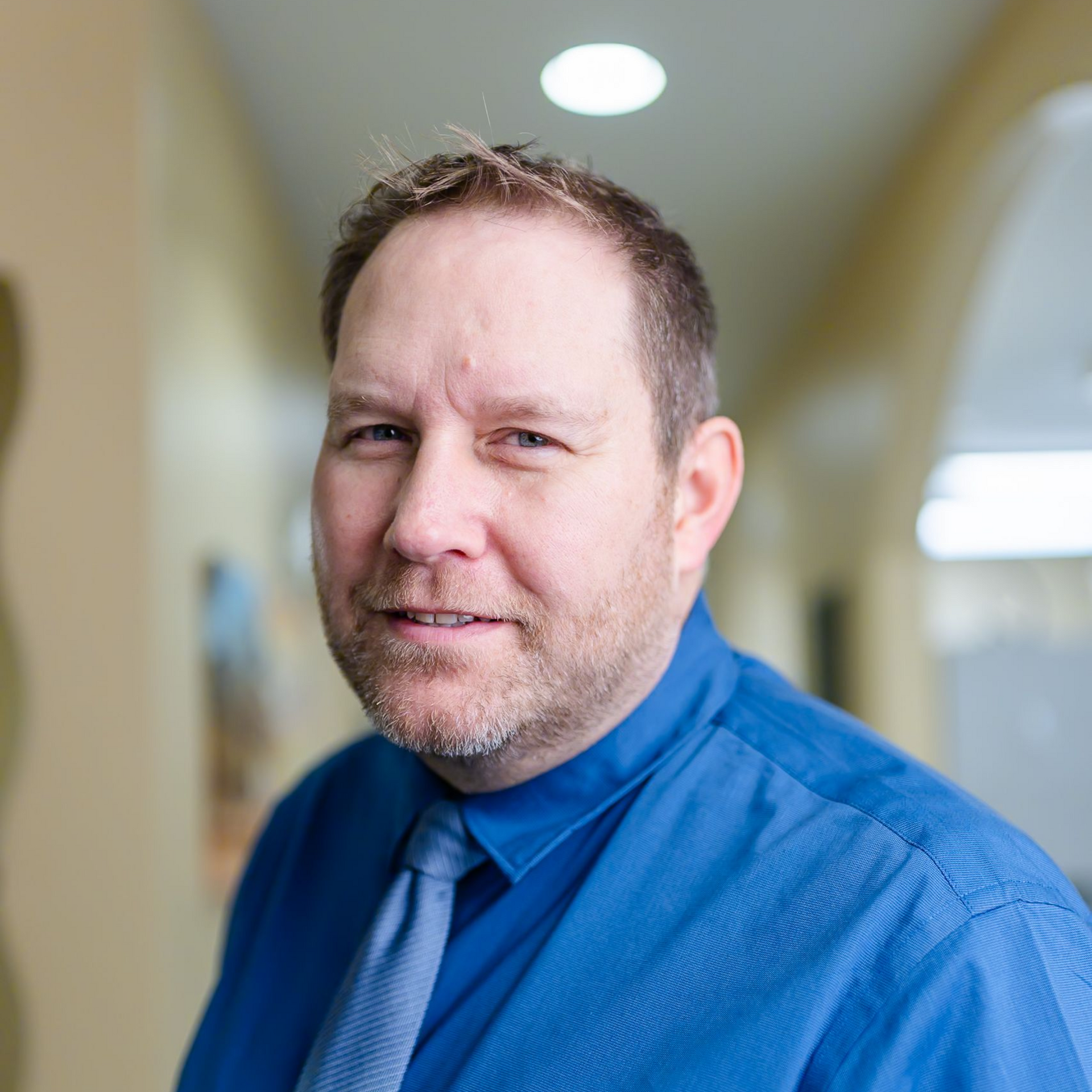 Man in a suit smiles, blue shirt and striped tie, against a blue background.