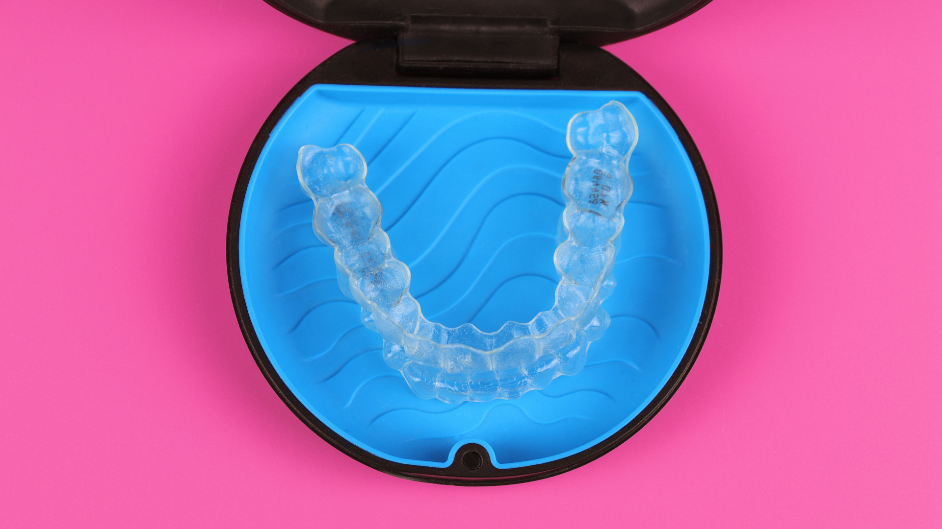Clear dental aligner in a blue case on a pink background.