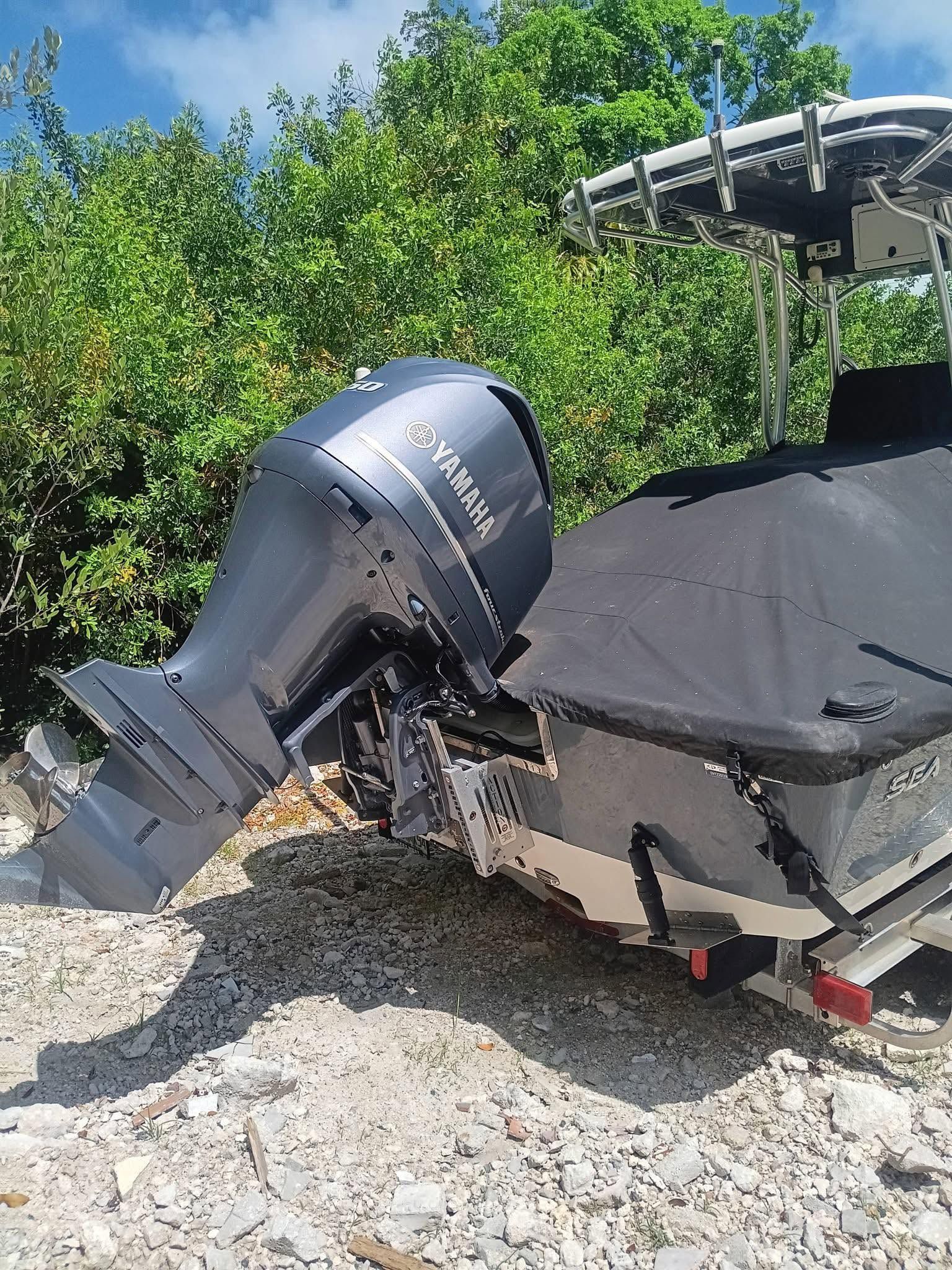 Broken boat motor leaning on damaged covered boat in gravel yard, with trees in the background