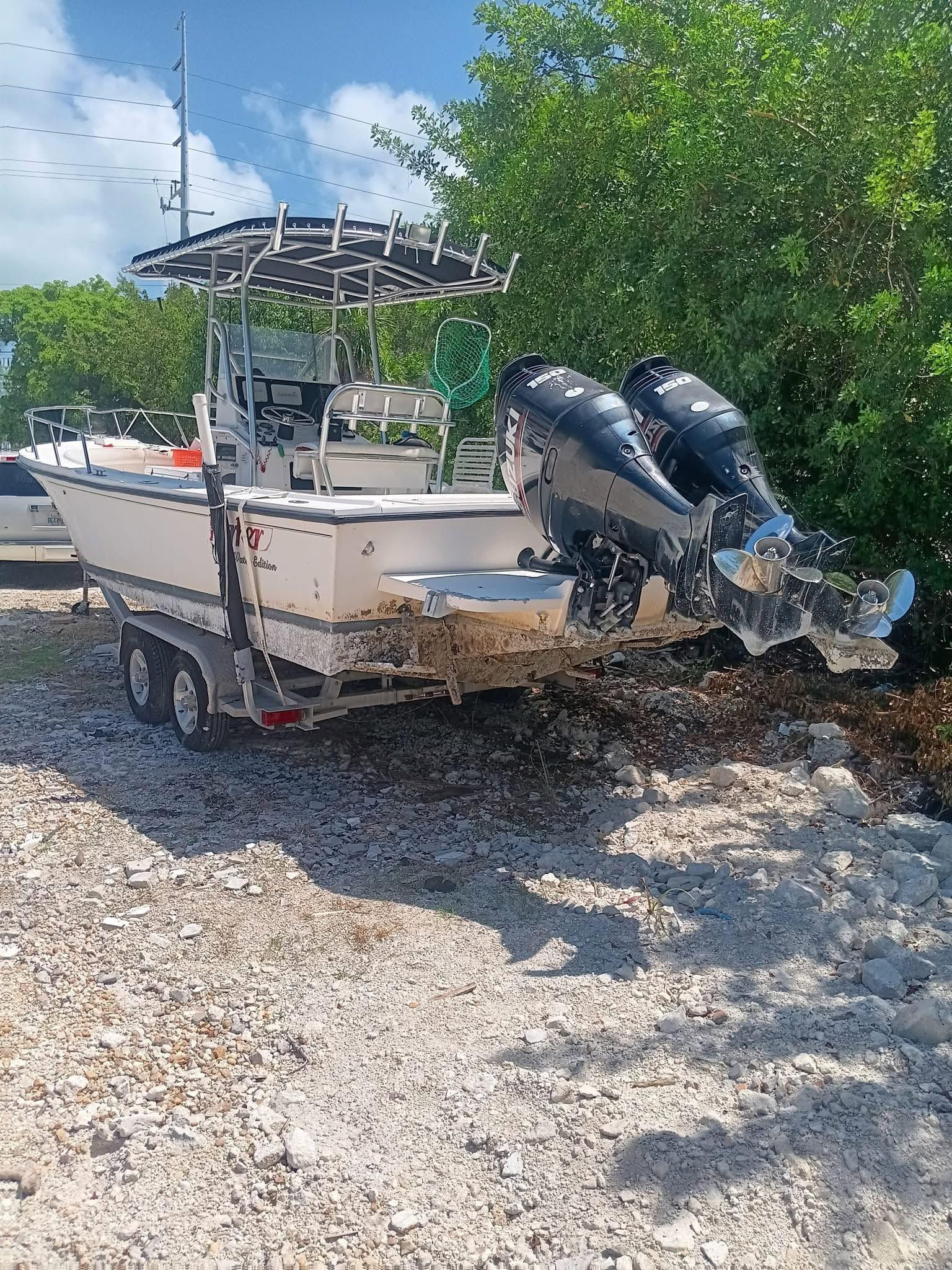 White fishing boat with twin outboard motors on a trailer in a gravel lot under a sunny sky