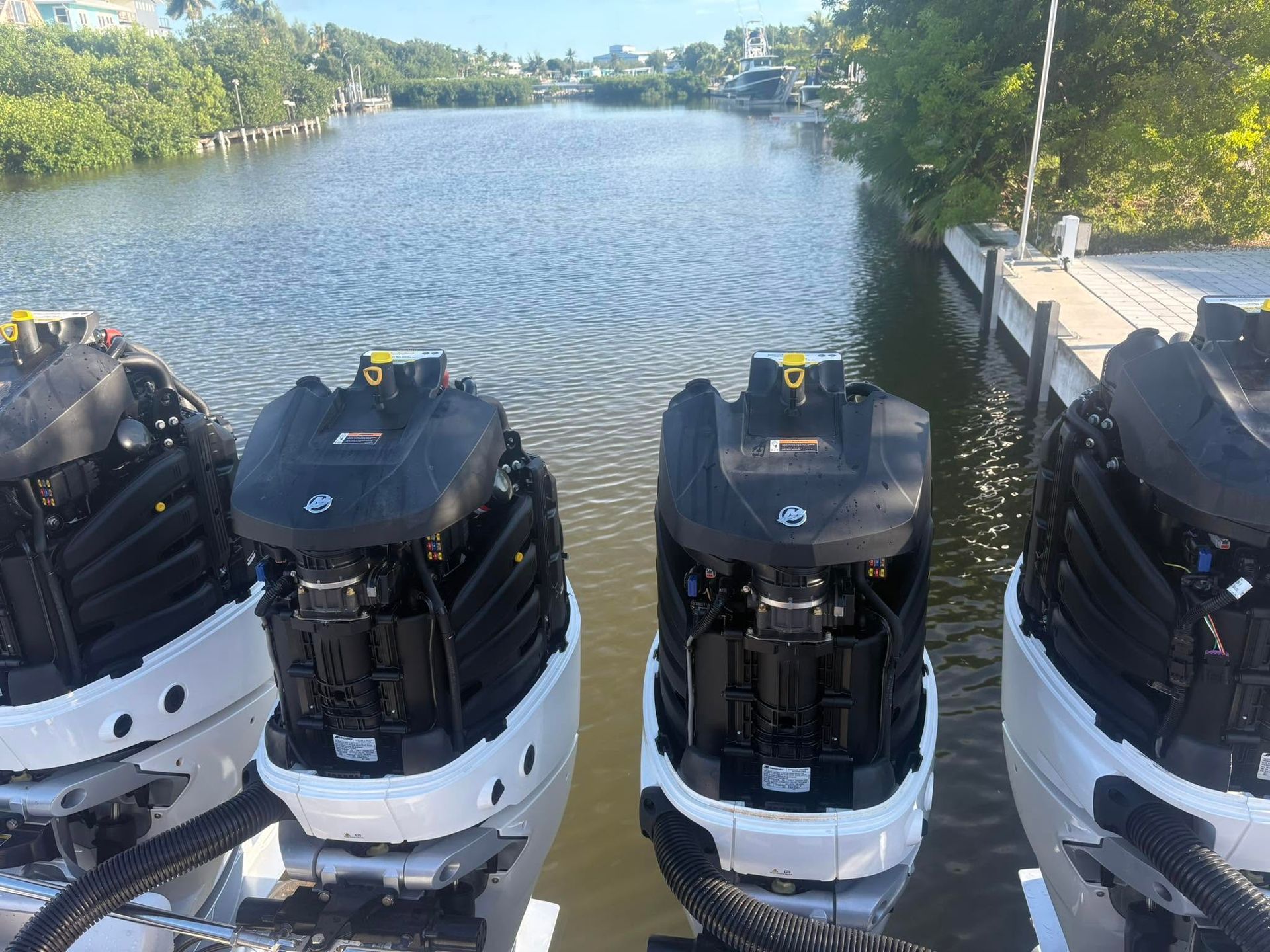 Four outboard boat motors lined up on a dock beside a calm canal under a sunny sky
