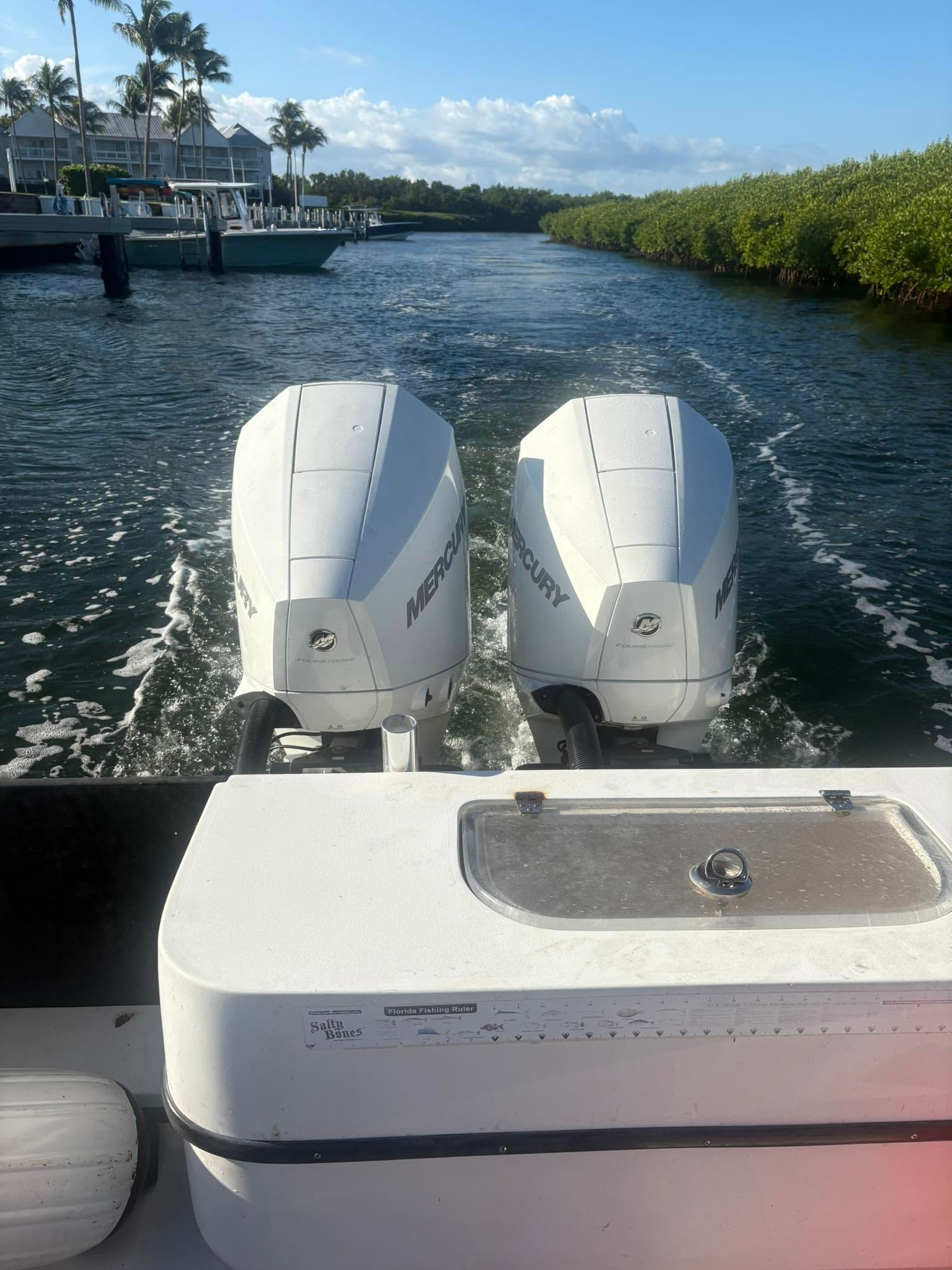 View from a boat: twin white outboard motors on a sunny canal with mangroves and waterfront homes.