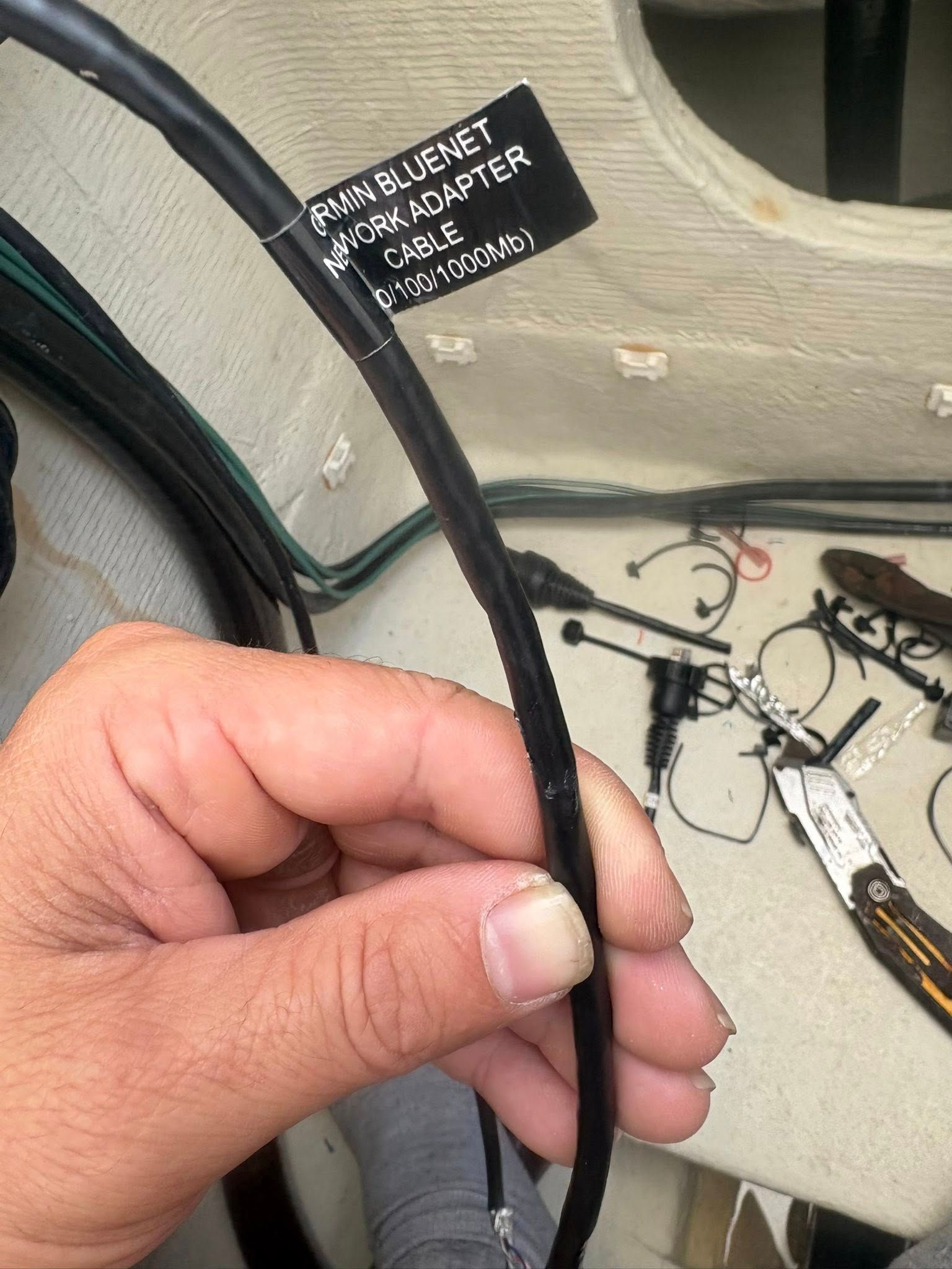 Hand holding a black cable with a white label near a beige wall and scattered wires.