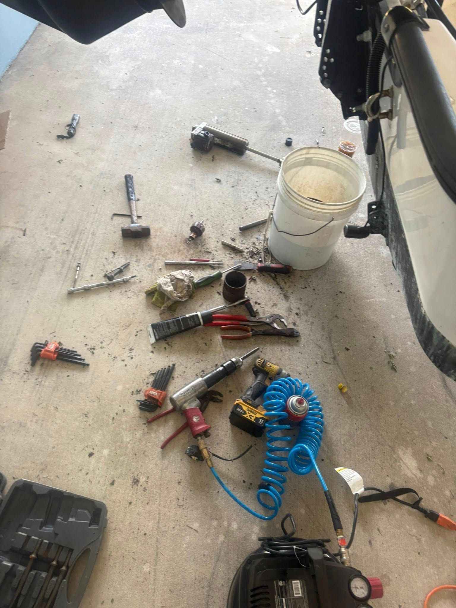 Scattered hand tools and paint supplies on a concrete garage floor next to a white bucket and car tire