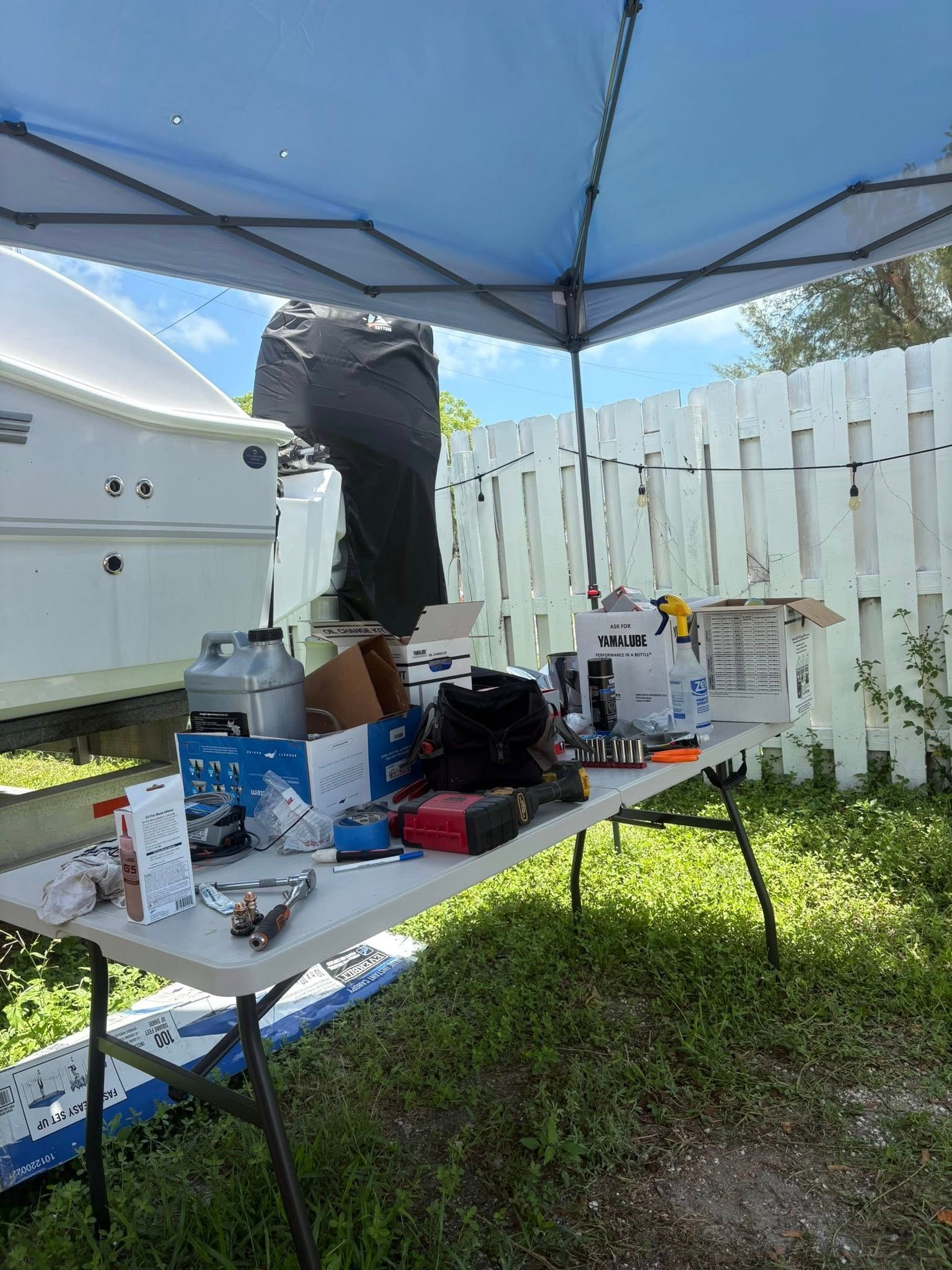Outdoor table under blue canopy with assorted tools and supplies beside a white fence