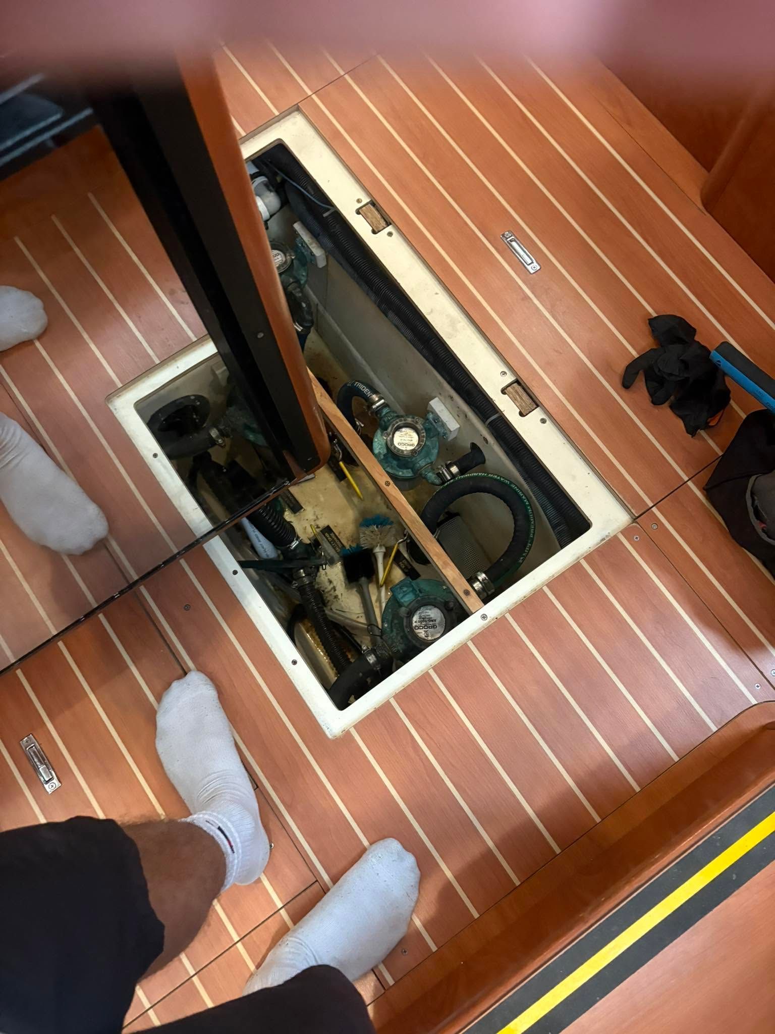View through a glass floor panel into a ship’s engine room, with two people standing on the wooden deck above.