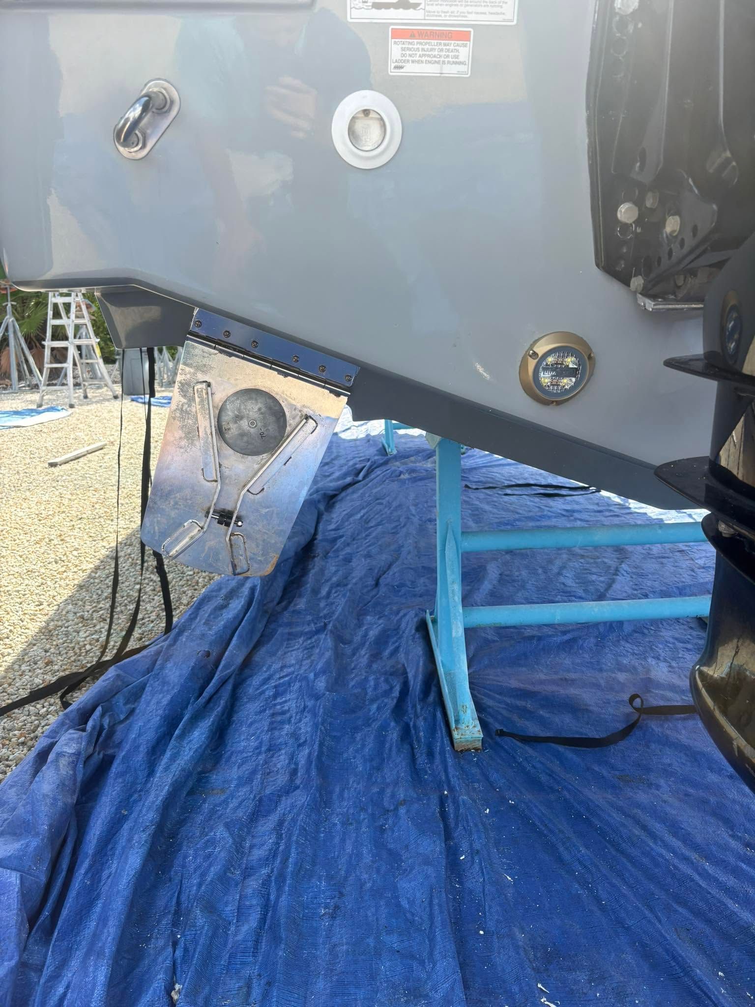 Underboat view of a boat hull on a blue tarp with support stands and a gravel lot beneath