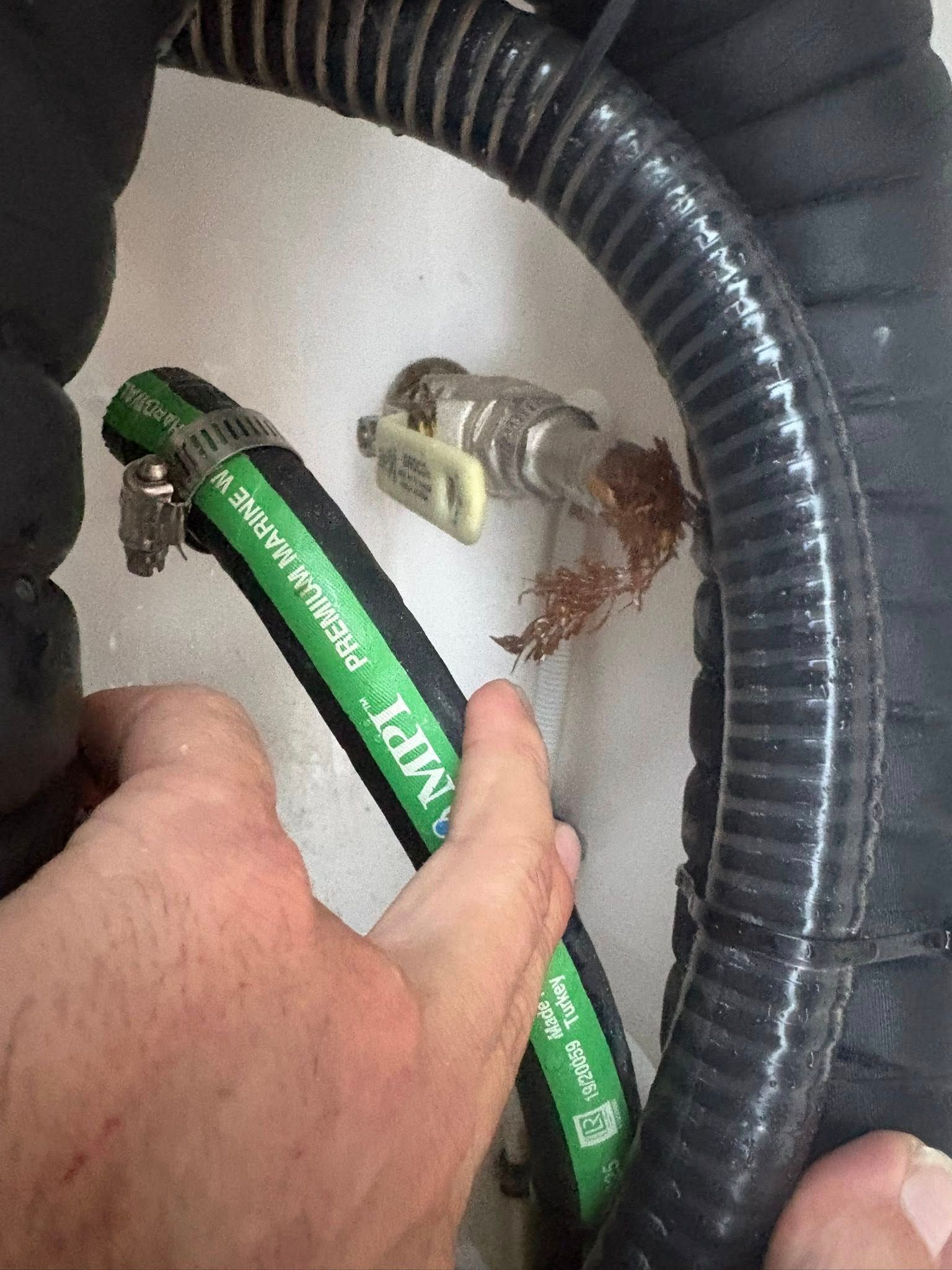 Hand holding a green hose near a metal fitting with a corroded connection inside a black corrugated tube