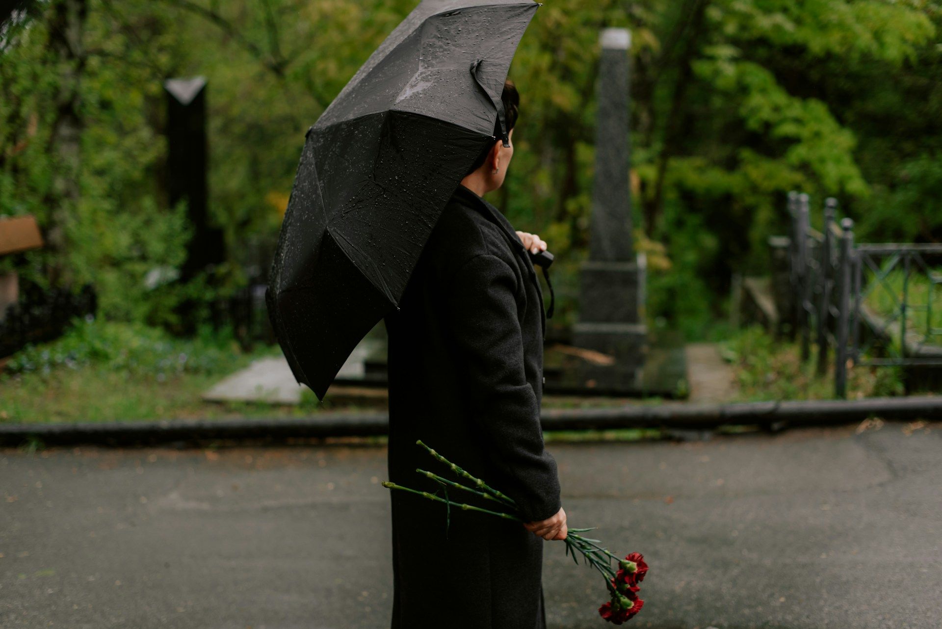 A person in a black coat holds a black umbrella and a bunch of red carnations in a cemetery on a rainy day.