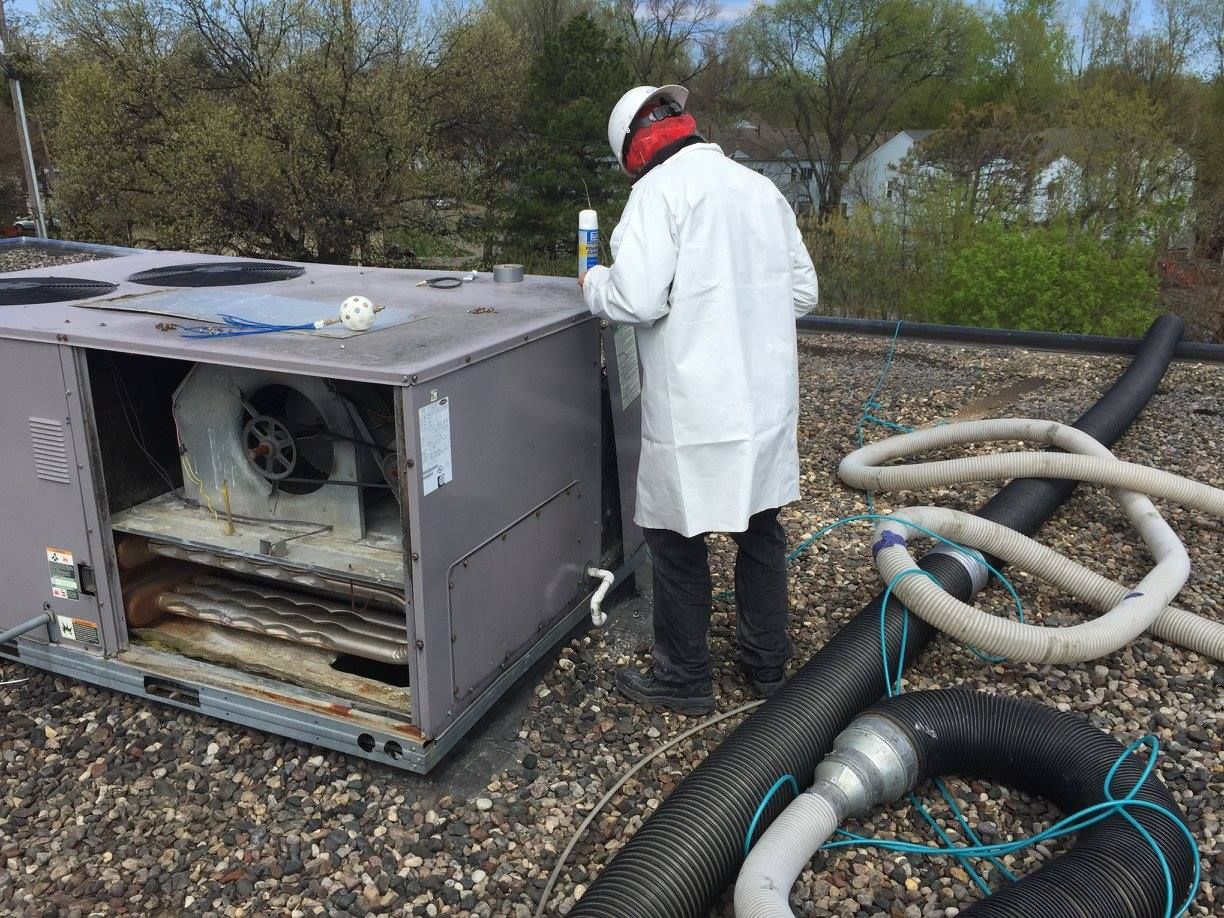 Person in a lab coat inspecting an HVAC unit on a rooftop. Air hoses are nearby.