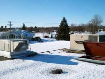 Snow-covered rooftop with HVAC units against a blue sky, trees, and buildings in the distance.