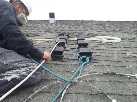 Roofer in hardhat and respirator, working on a shingled roof, securing equipment with ropes.