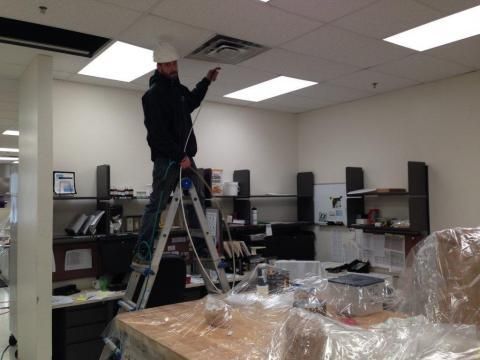 Man on ladder works on ceiling fixture in office with cluttered desks.
