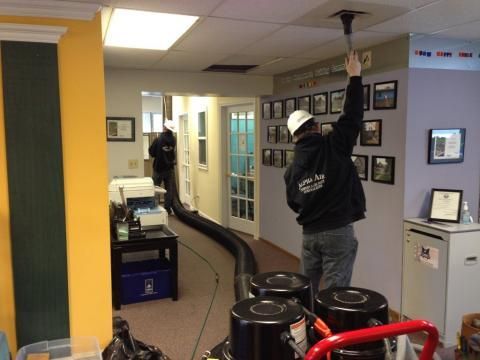 Workers vacuuming ceiling vents in an office, using large industrial equipment.