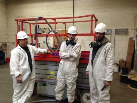 Three people in white protective suits and hard hats stand near a red lift in an indoor workspace.