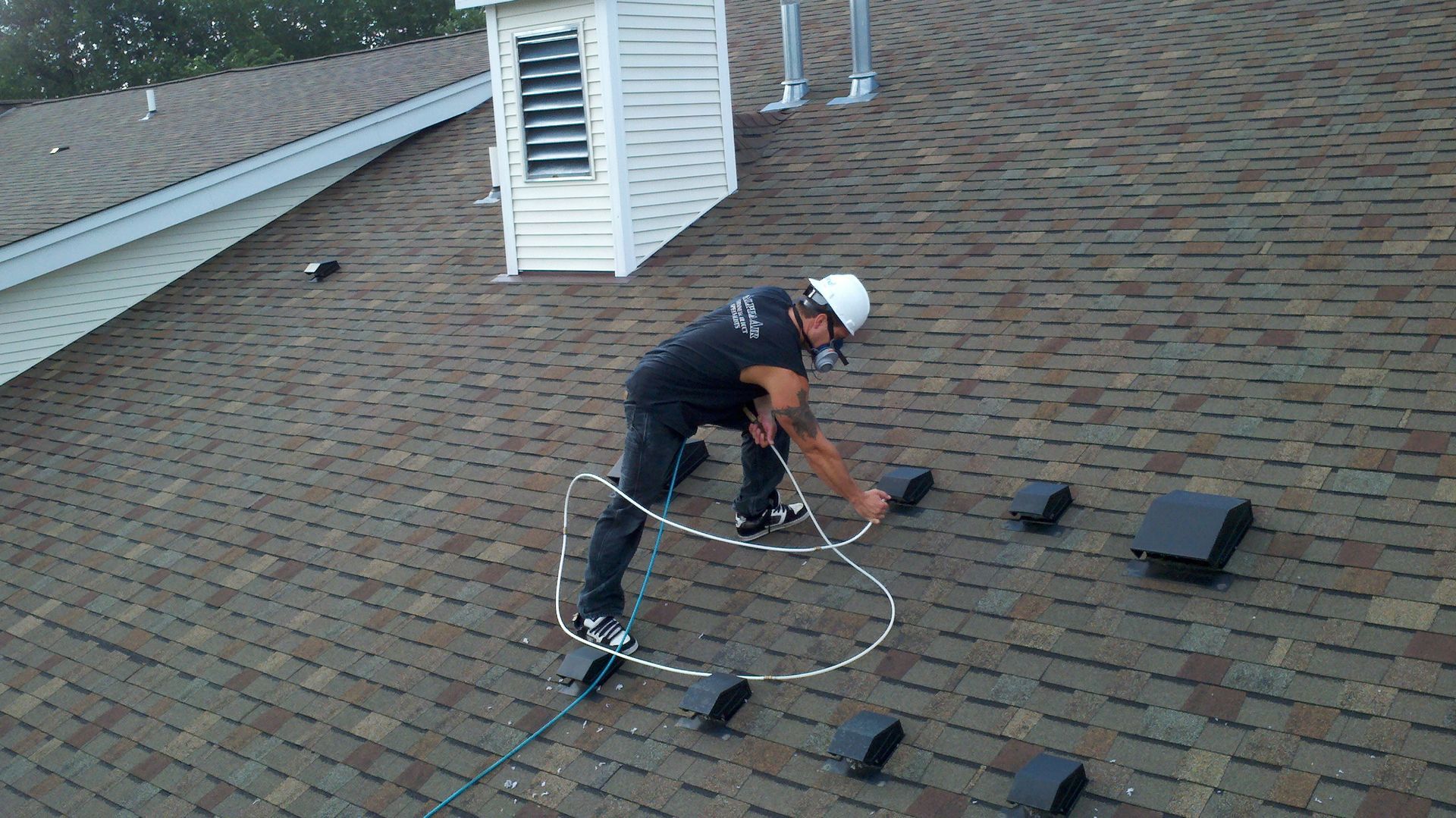 Alpha Air Corporation technician on a brown shingled roof, wearing a hard hat, inspecting with a tool.