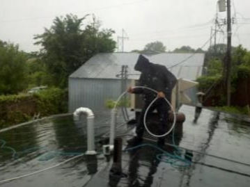 Person in rain gear on a wet roof, working with a hose. Buildings and trees in background.