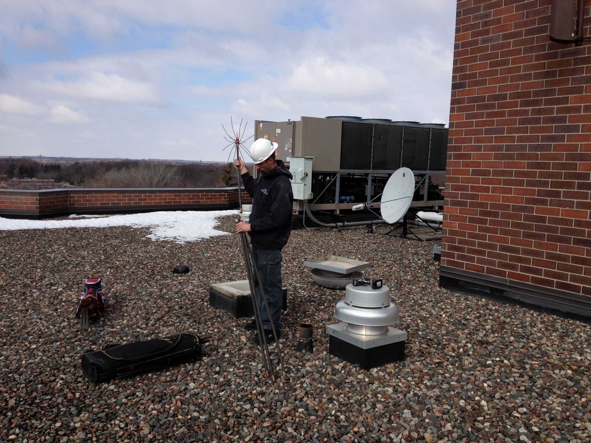 Man in hard hat on rooftop, holding device, near HVAC unit and antennas.