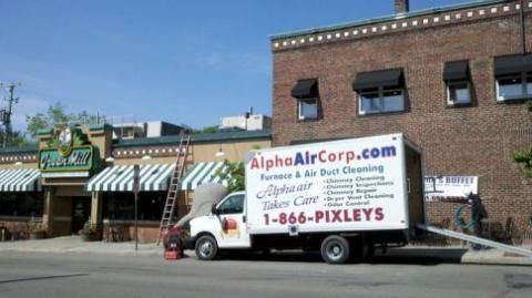 White Alpha Air Corp truck parked in front of a brick building and a restaurant; the truck's ramp is down.