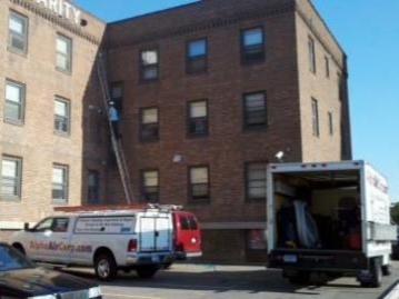Exterior view of a brick building with a ladder against it, a worker on the ladder, and moving trucks.