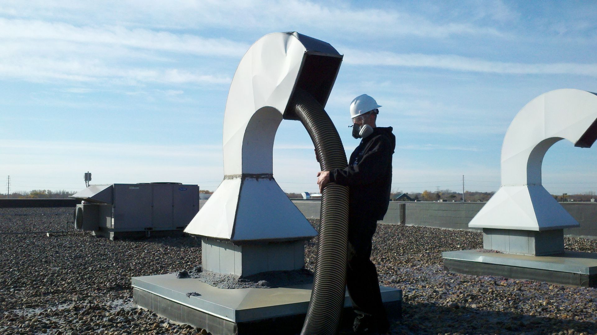 Man in hard hat holds a large, dark flexible pipe attached to a roof vent on a flat rooftop.