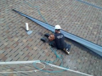 Roofer on a shingled roof, cutting a piece of metal flashing with a power tool.
