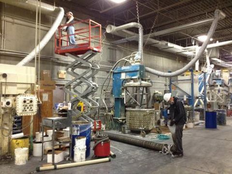 Two workers near industrial machinery, one in a lift, the other on the ground, inside a large warehouse.