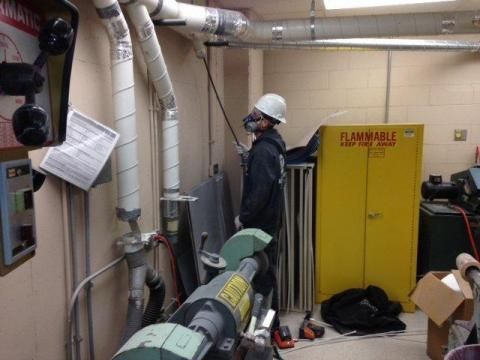 Person in hard hat and respirator sprays overhead ductwork in a workshop with a yellow flammable cabinet.