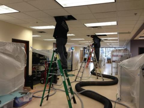 Two workers on ladders accessing ceiling vents in a room with covered equipment; long black hoses run along the floor.
