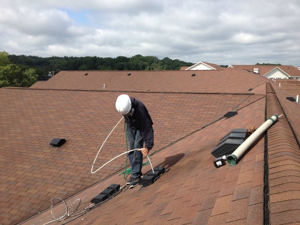 Roofer on a brown shingled roof, installing equipment, overcast sky.